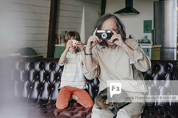 Großvater mit Vintage-Kamera und Enkelin mit Spielzeugkamera fotografieren zu Hause
