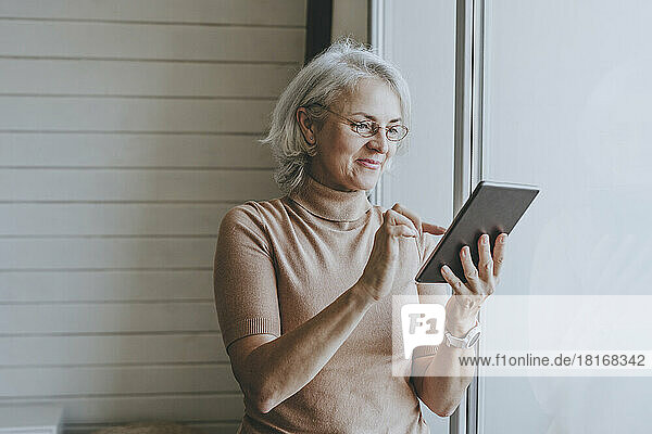 Smiling woman using tablet PC standing by window at home