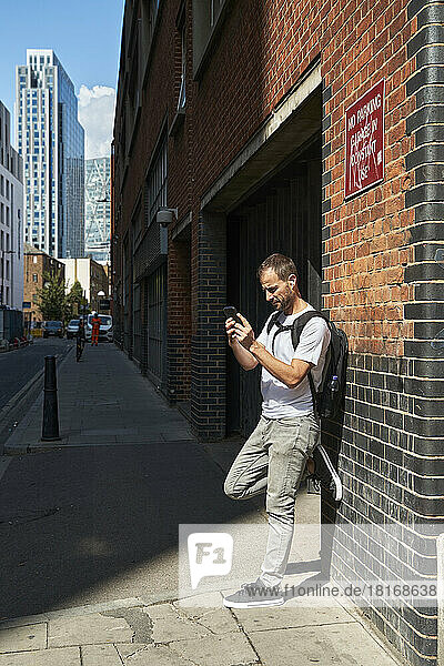 Man using smart phone leaning on wall at footpath