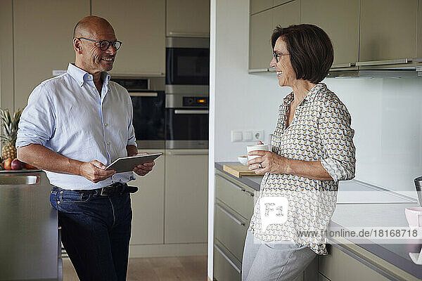 Happy senior man holding tablet PC standing with woman in kitchen at home
