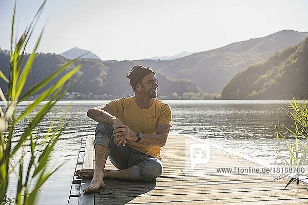 Smiling man with knit hat sitting on jetty over lake