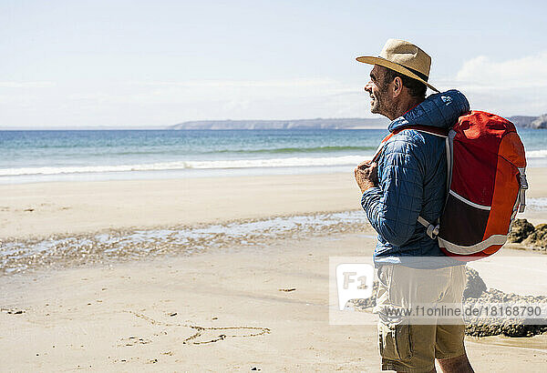 Mann mit Rucksack und Hut steht an einem sonnigen Tag am Strand