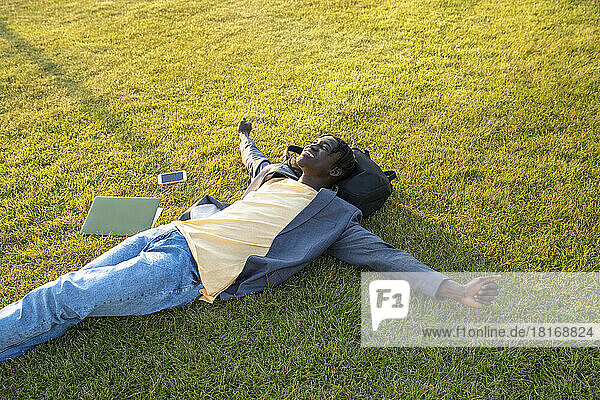 Smiling businessman with mobile phone and laptop relaxing on grass