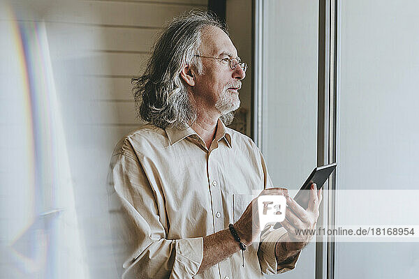 Nachdenklicher älterer Mann mit Tablet-PC am Fenster zu Hause