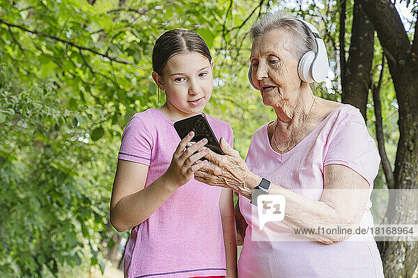 Granddaughter helping great grandmother using smartphone and headphones at park