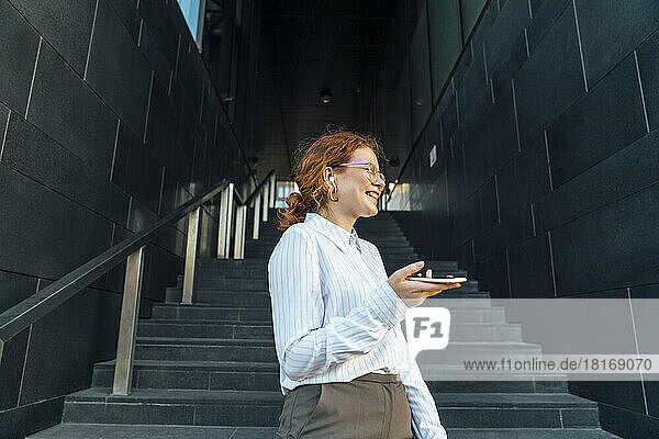Happy businesswoman with smart phone talking through wireless in-ear headphones