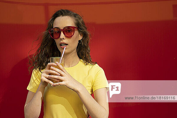 Woman wearing sunglasses holding juice glass in front of red wall