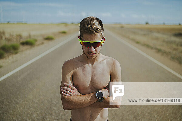 Shirtless young man wearing sunglasses standing with arms crossed on road