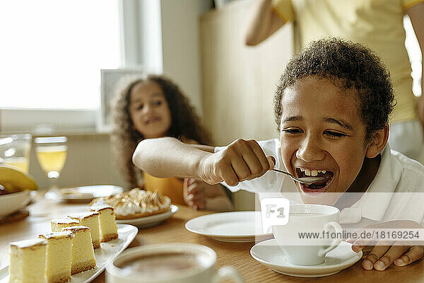 Glücklicher Junge trinkt heiße Schokolade mit Löffel von Schwester am Esstisch