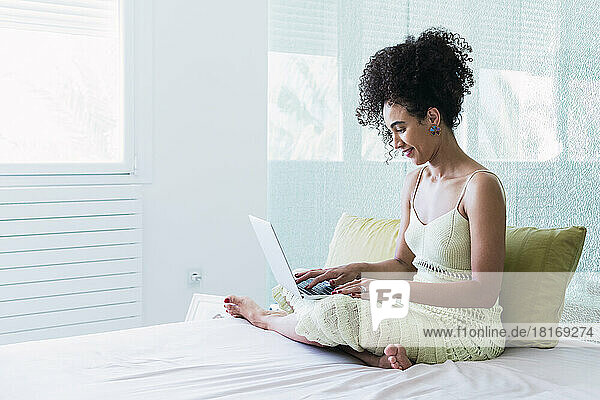 Smiling woman with curly hair using laptop sitting on bed at home