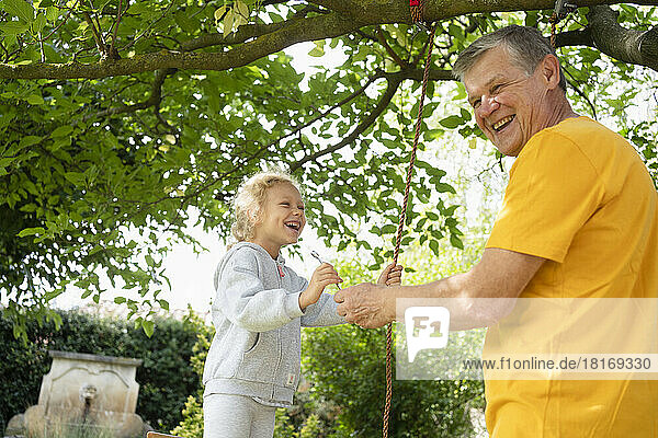 Glücklicher älterer Mann mit Enkelin beim Schaukeln mit Seil im Garten
