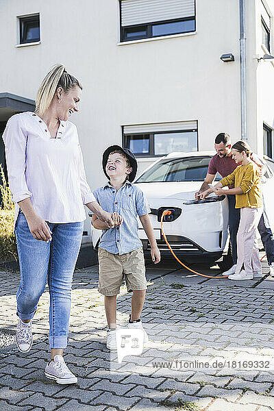 Happy mother looking at son while father and daughter are charging electric car