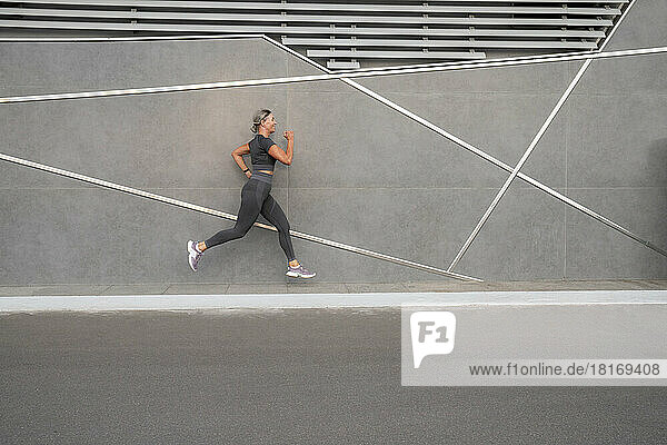 Mature woman jogging on footpath by gray wall