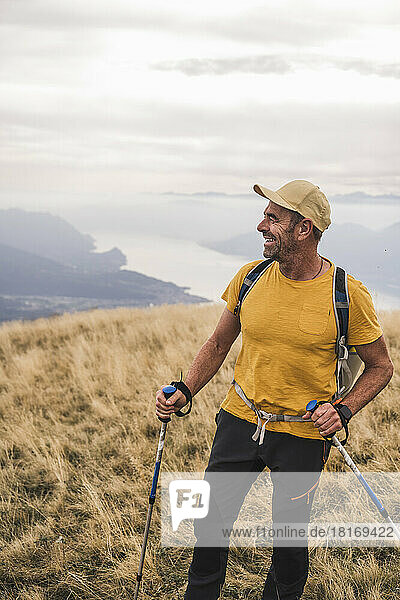Happy mature man wearing cap standing with hiking poles under cloudy sky