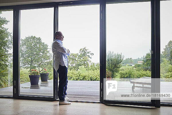Thoughtful senior man with arms crossed leaning on glass door at home