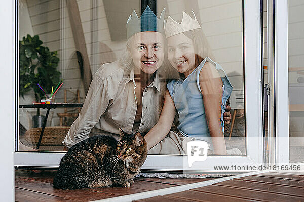 Glückliche Großmutter und Enkelin mit Krone sitzen zu Hause gemeinsam hinter einem Glasfenster