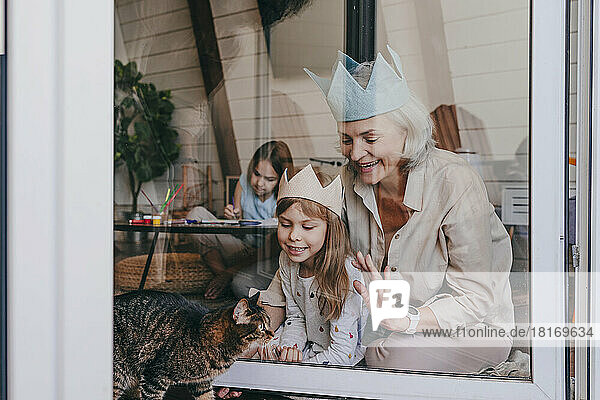 Großmutter und Enkelin tragen eine Krone und schauen durch ein Glasfenster auf die Katze