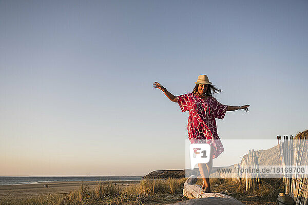 Happy mature woman with arms outstretched walking on log at beach