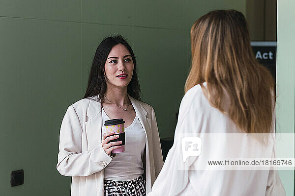 Businesswoman with disposable coffee cup looking at colleague in office