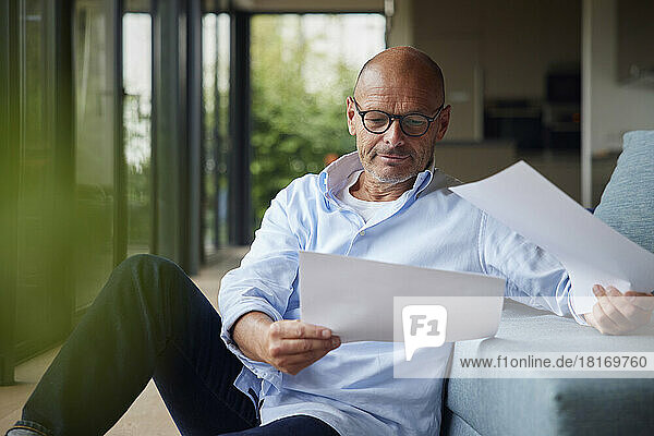 Senior man with documents sitting by sofa at home