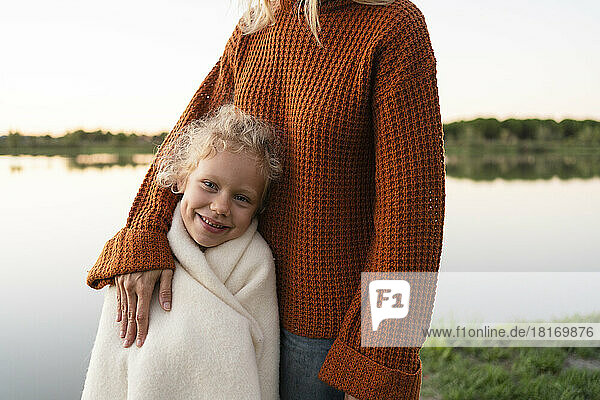 Smiling girl wrapped in blanket standing with mother at lake