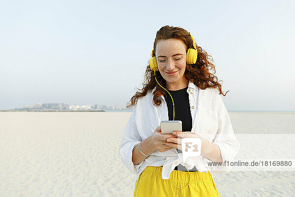 Smiling woman wearing headphones using smart phone at beach