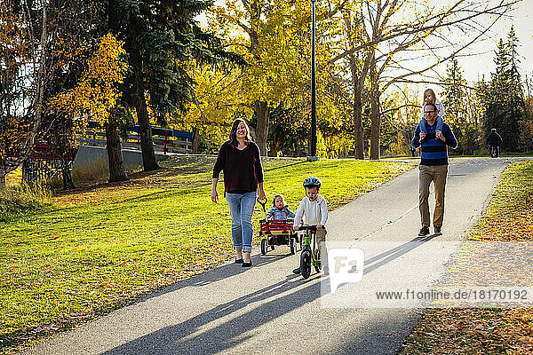 Eine Mutter zieht ihre Tochter mit Down-Syndrom in einem Wagen  der Vater hat die Tochter auf den Schultern und der Sohn fährt mit dem Fahrrad in einem Stadtpark während eines Familienausflugs in der Herbstzeit; St. Albert  Alberta  Kanada
