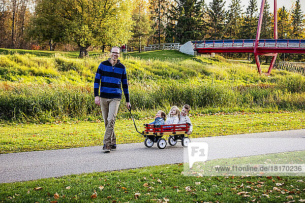 A father pulling his young children in a wagon along a river in a city park with a bridge in the background during the fall season and his baby girl has Down syndrome; St. Albert  Alberta  Canada