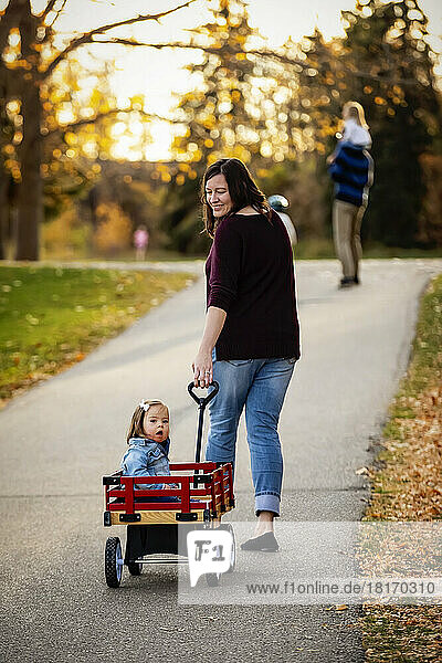 Eine Mutter zieht ihr Baby mit Down-Syndrom in einem Wagen entlang eines Flusses in einem Stadtpark mit ihrer Familie während der Herbstsaison; St. Albert  Alberta  Kanada