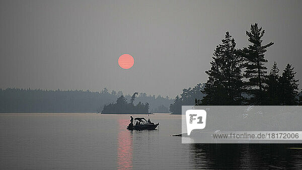 Bootfahren und Angeln auf einem See bei Sonnenuntergang. Die rosafarbene Sonne leuchtet über einer silhouettierten Landschaft und Wasser mit einem rosafarbenen Sonnenstrahl  der auf das Wasser scheint  Lake of the Woods  Ontario; Kenora  Ontario  Kanada