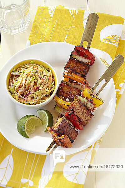 Grilled tofu and bell peppers on bamboo skewers with side dish of slaw and lime wedges on a yellow patterned napkin