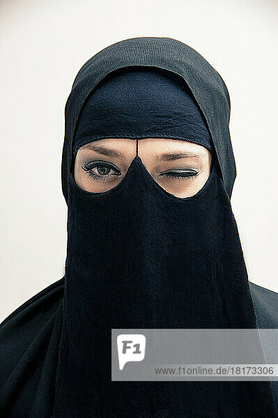 Close-up portrait of young woman wearing black  muslim hijab and muslim dress  winking and looking at camera  eyes showing eye makeup  studio shot on white background