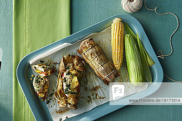 Chicken breast stuffed with red peppers  herbs and corn wrapped in a corn husk and cooked on the barbeque. Shown with a raw cob of corn and twine to tie the husks on a tabletop.