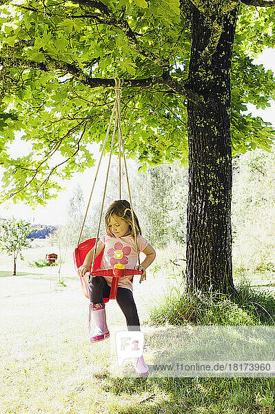 3 year old girl in rubber boots sitting in red swing in back yard  Sweden