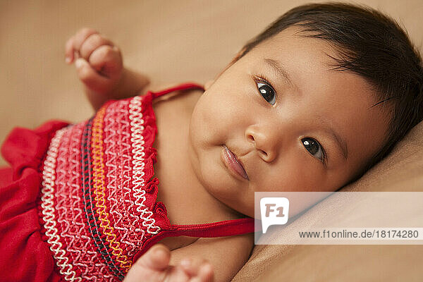 Close-up portrait of Asian baby lying on back  wearing red dress  looking at camera  studio shot on brown background