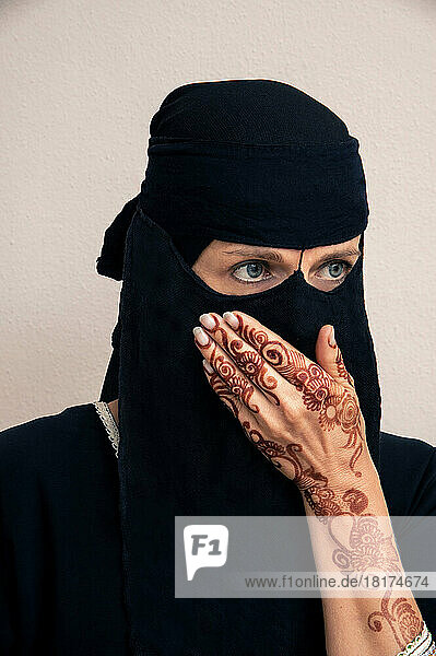 Close-up portrait of woman wearing black muslim hijab and muslim dress  looking to the side with hand covering mouth and showing arms and hands painted with henna in arabic style  studio shot on whtie background