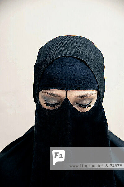 Close-up portrait of young woman wearing black  muslim hijab and muslim dress  eyes closed showing eye makeup  studio shot on white background