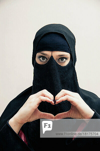 Close-up portrait of young woman wearing black  muslim hijab and muslim dress  making heart shape with hands  looking at camera  eyes showing eye makeup  studio shot on white background