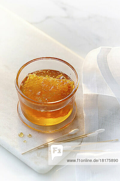 A small glass jar with clear golden honey and a honey comb with cotton swabs. High key image of honey and beauty products.