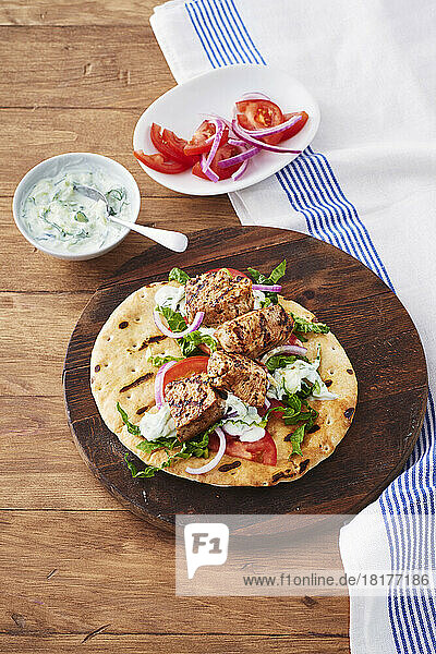 Pork souvlaki on a grilled pita on a wooden cutting board with side dishes of tzatziki and chopped tomatoes and sliced onions