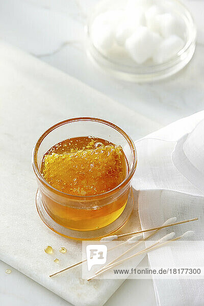 A small glass jar with clear golden honey and a honey comb with cotton swabs and cotton balls. High key image of honey and beauty products.
