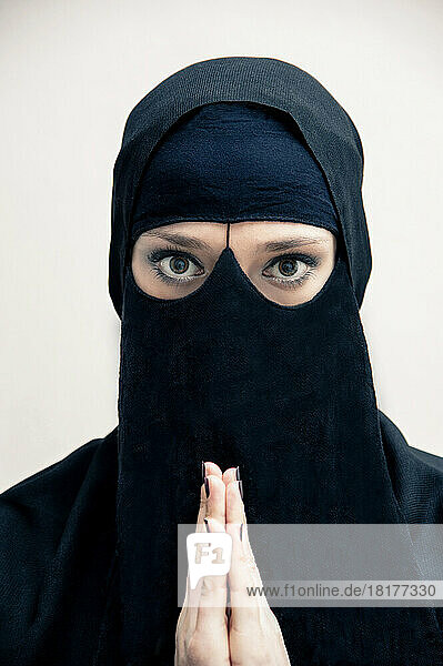 Close-up portrait of young woman wearing black  muslim hijab and muslim dress  with hands praying  looking at camera  eyes showing eye makeup  studio shot on white background