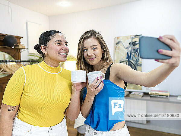 Smiling woman holding coffee cup taking selfie with friend through mobile phone