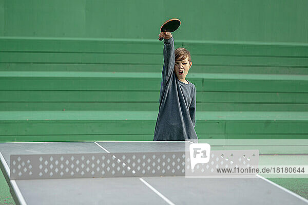 Boy holding table tennis racket screaming in front of green wall