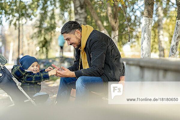 Happy baby boy in stroller holding hands of father sitting under tree at park