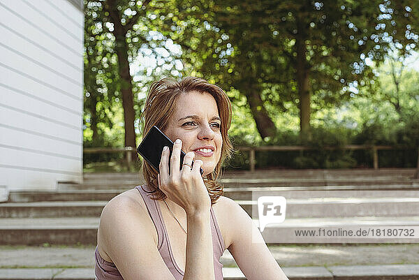 Lächelnde Frau  die auf einer Treppe im Park sitzt und mit ihrem Smartphone spricht