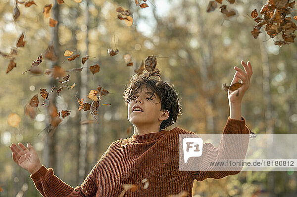 Verspielter Junge mit geschlossenen Augen wirft trockene Blätter in den Park