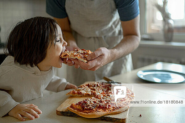 Man feeding pizza to son in kitchen at home