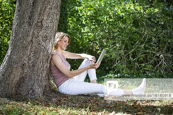 Glückliche Frau mit Tablet-PC sitzt am Baumstamm im Park