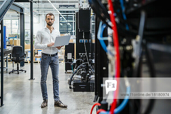 Businessman with laptop standing near machinery in industry